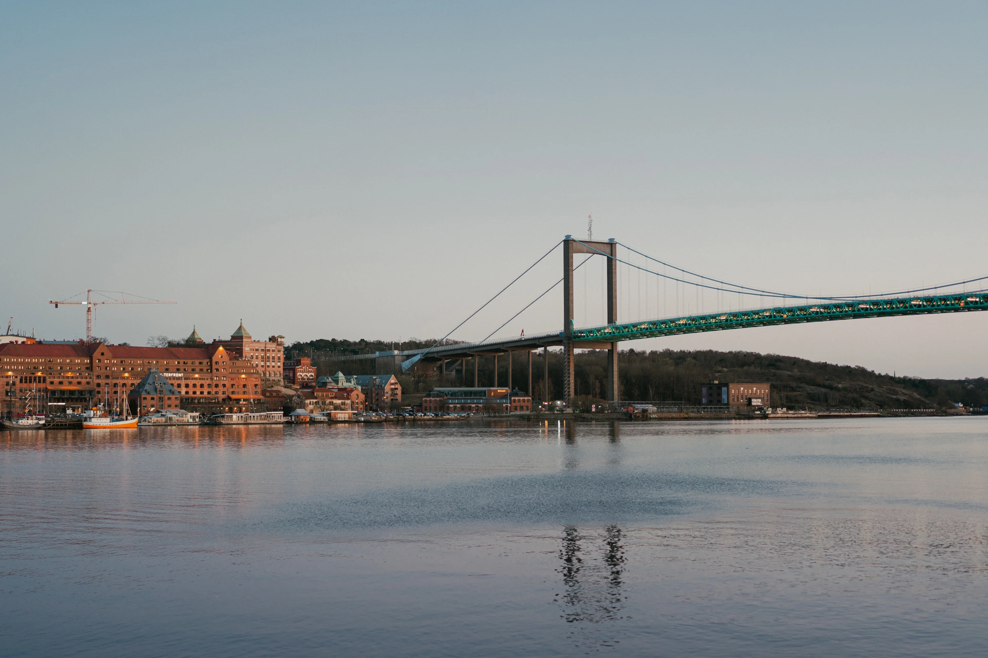 The teal Älvsborg suspension bridge over calm water at dusk in Gothenburg, with a red-brick waterfront and a moored boat on the left