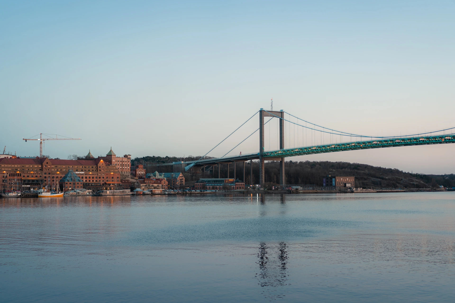 The teal Älvsborg suspension bridge over calm water at dusk in Gothenburg, with a red-brick waterfront and a moored boat on the left