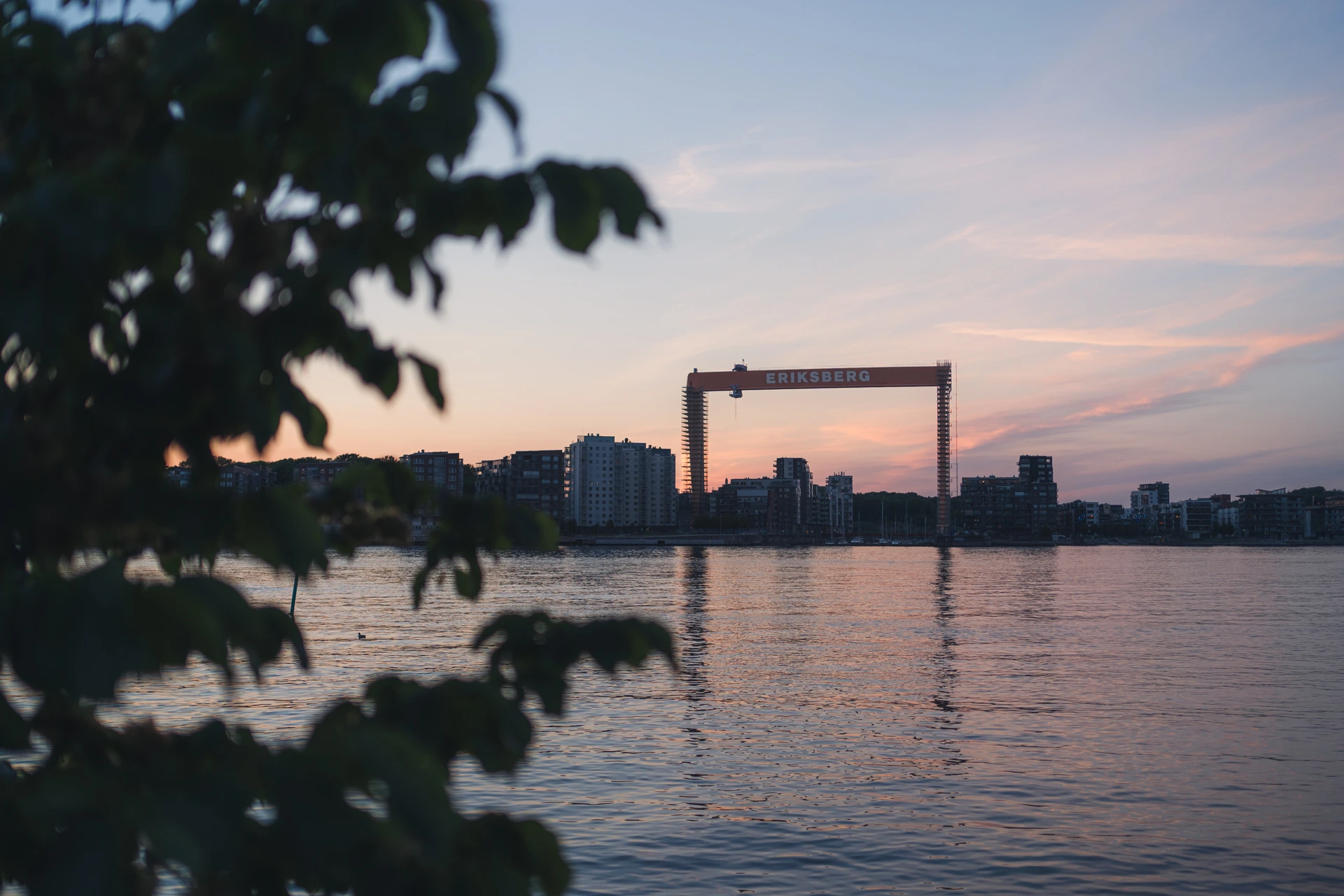 The red Eriksberg shipyard gantry crane on the Gothenburg waterfront at sunset, seen across the river through out-of-focus leaves