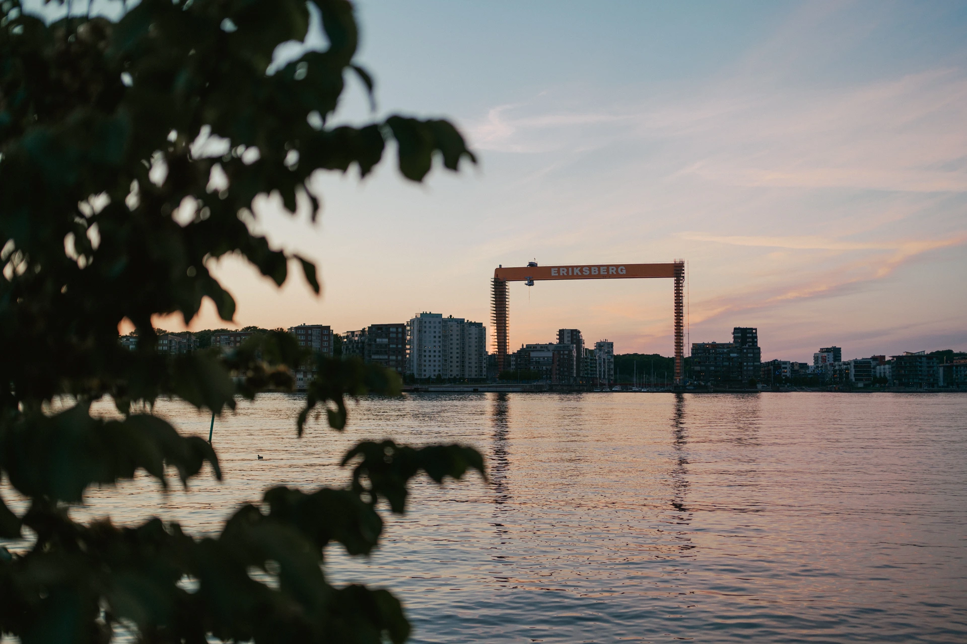 The red Eriksberg shipyard gantry crane on the Gothenburg waterfront at sunset, seen across the river through out-of-focus leaves