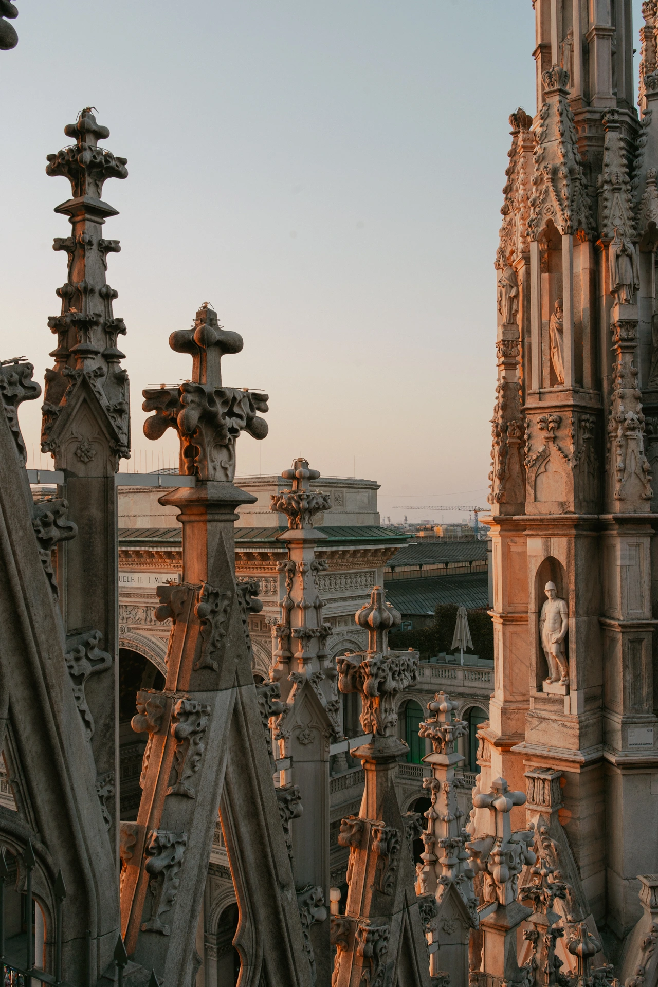 Carved marble spires and statues on the roof of the Duomo in Milan at sunset, the stone lit warm orange, with the facade of the Galleria Vittorio Emanuele II and the city rooftops visible between them