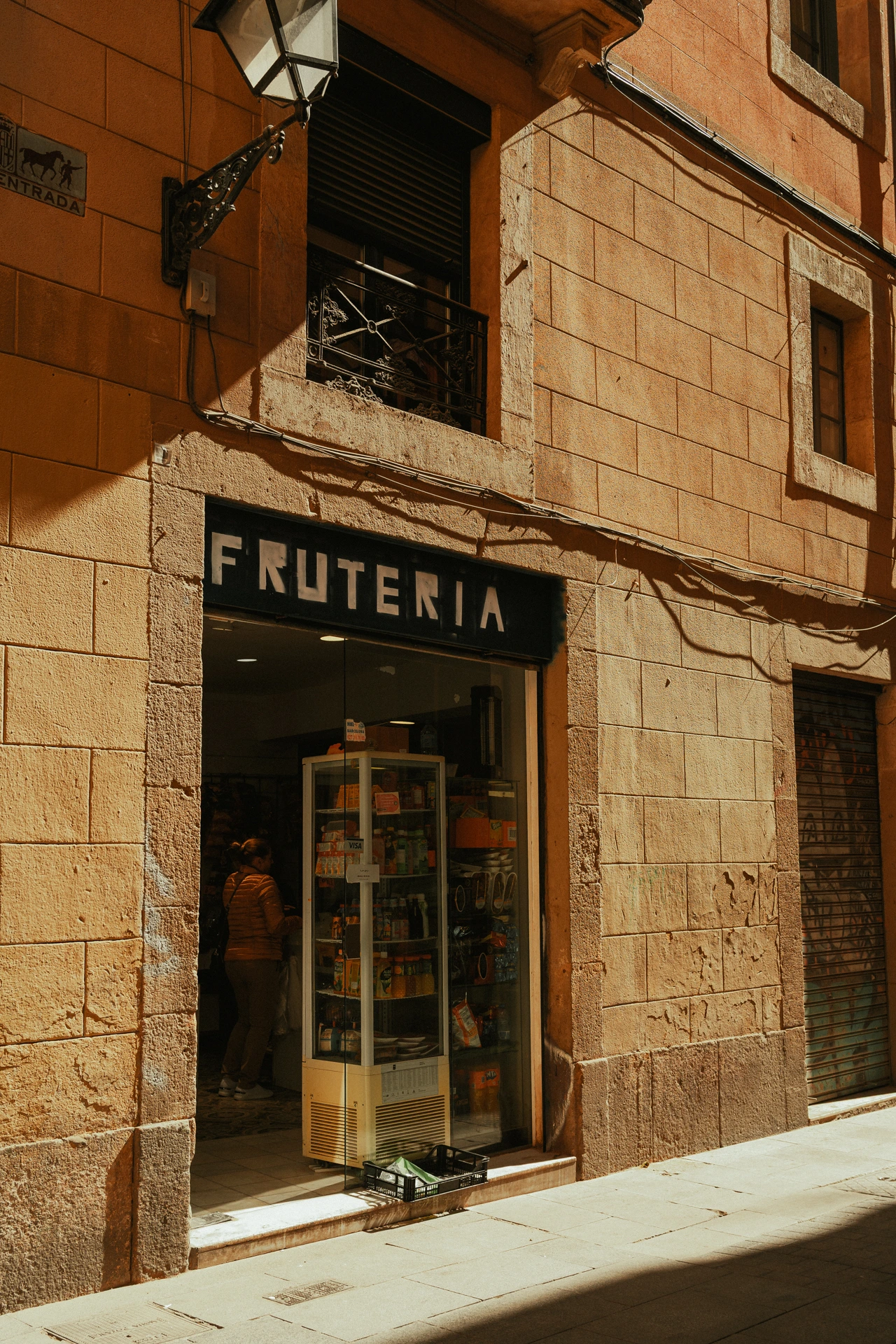 A small frutería in the old town of Barcelona, its black shopfront lettered in white above the open doorway, late afternoon sun raking across the warm sandstone facade and a wrought-iron street lantern, with a customer standing in the shaded interior beside a glass drinks cooler