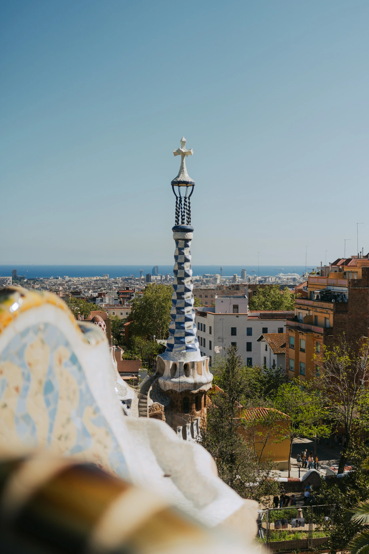 The mosaic-tiled spire of a Park Güell gatehouse in Barcelona under a clear blue sky, topped with a cross, overlooking the city rooftops stretching to the Mediterranean sea in the distance