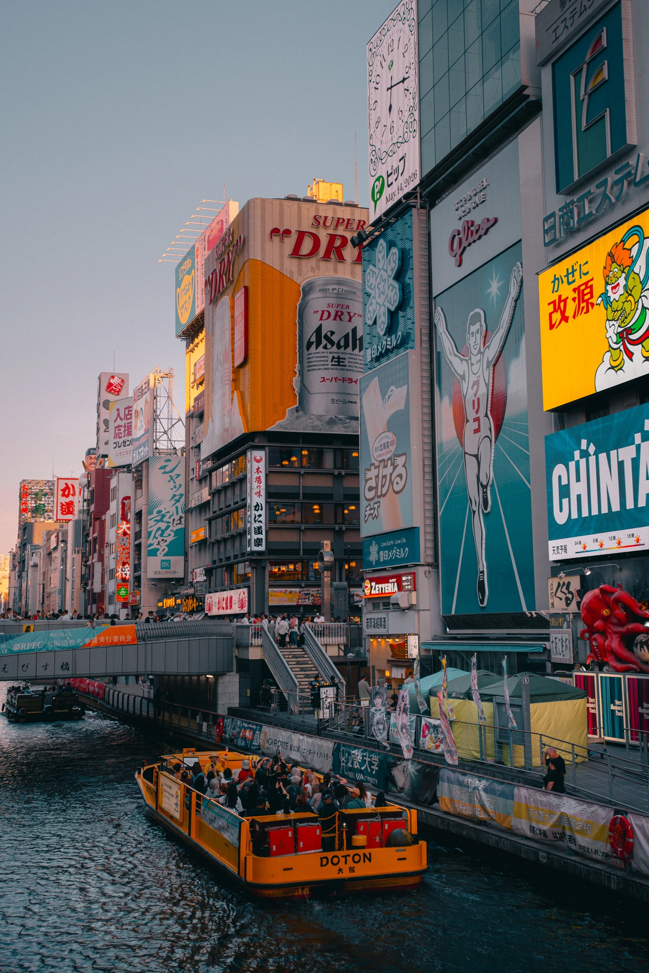 The Dotonbori canal in Osaka at dusk, a bright yellow open-top tour boat crowded with passengers passing below the wall of neon billboards along the waterfront, the Glico running man and a giant Asahi beer can among them, with people watching from the Ebisu bridge on the left