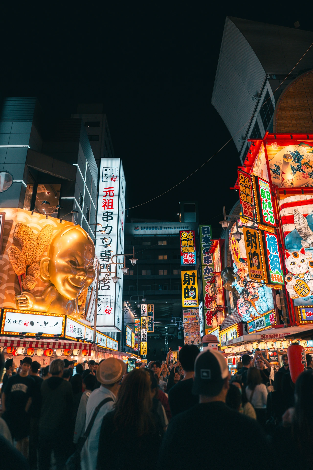 The Shinsekai district in Osaka at night, a crowd filling the street beneath a dense cluster of glowing neon signs and paper lanterns, with the giant golden Kushikatsu Daruma chef figure lit up on the left